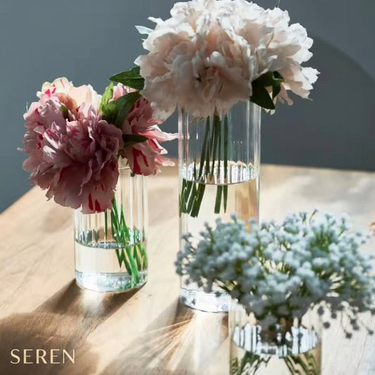 Three clear glass cylindrical vases with flared bases holding water and flowers, arranged on a light wood tabletop in natural sunlight. The vases contain pink peonies, a large white peony, and small white baby's breath.