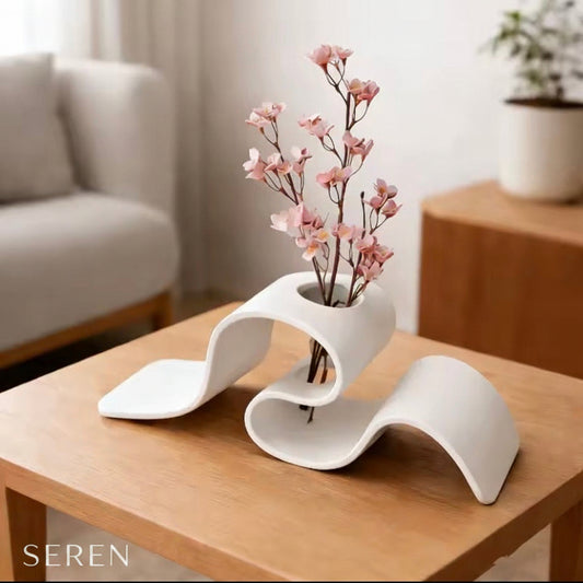 Modern white vase with pink flowers on a wooden table in a living room setting.