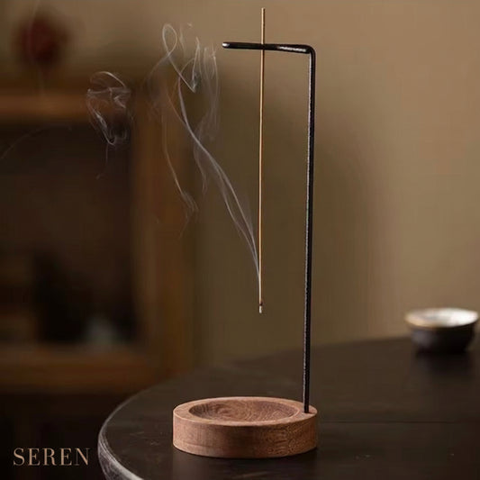 A black metal incense holder with a vertical stand and curved top bar, holding a single incense stick that is smoking upward. The holder sits on a round wooden base on a dark table, with a softly blurred indoor background.