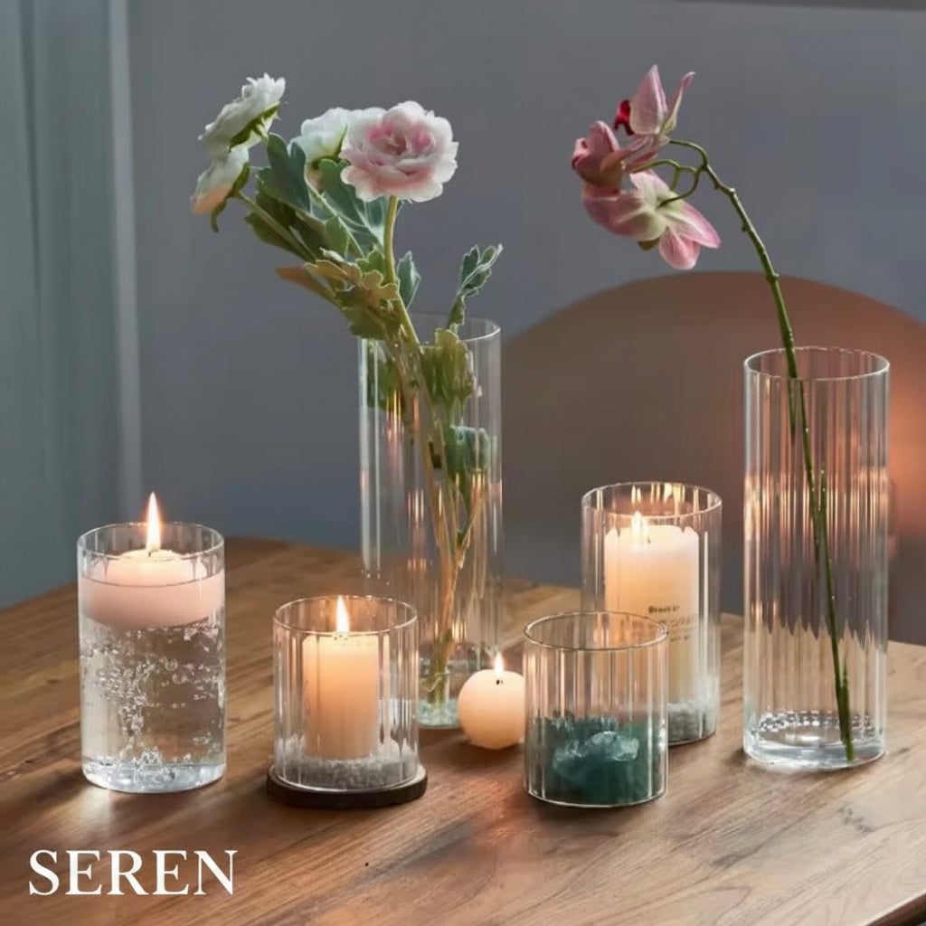 A styled arrangement of clear glass candle holders and tall fluted vases on a wooden table, with several lit candles and flowers including white and pink blossoms.