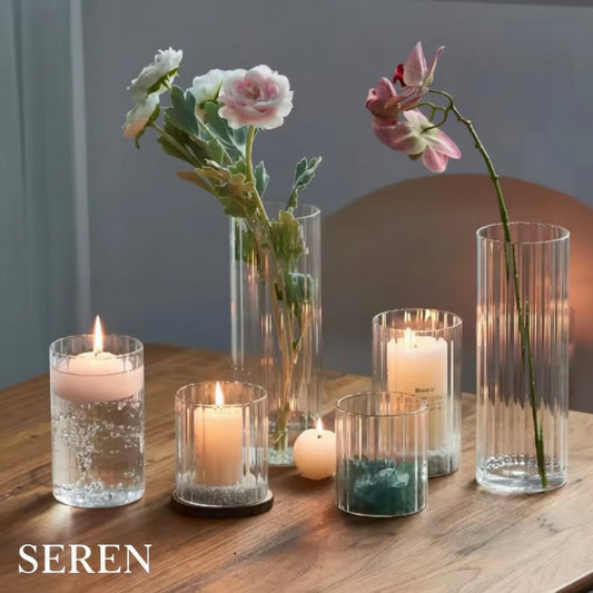 A styled arrangement of clear glass candle holders and tall fluted vases on a wooden table, with several lit candles and flowers including white and pink blossoms.