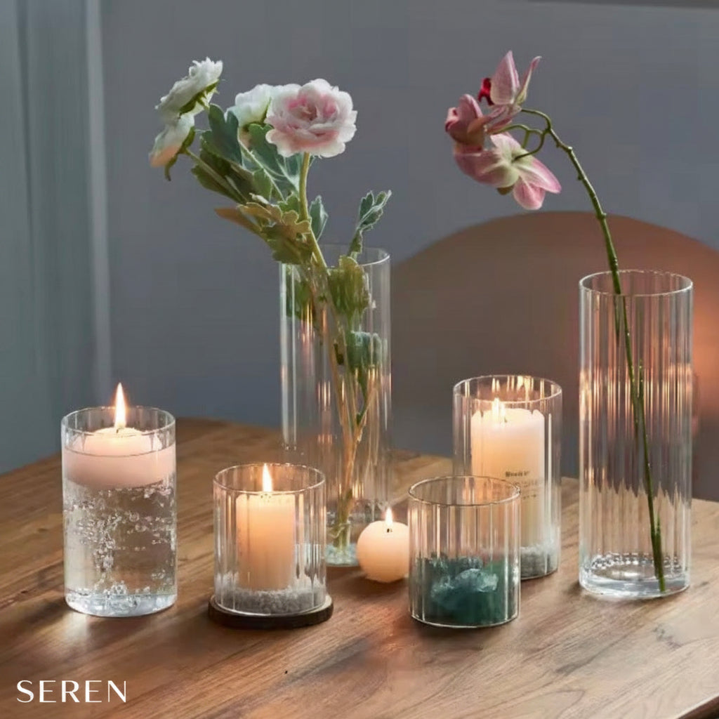 A styled arrangement of clear glass candle holders and tall fluted vases on a wooden table, with several lit candles and flowers including white and pink blossoms.