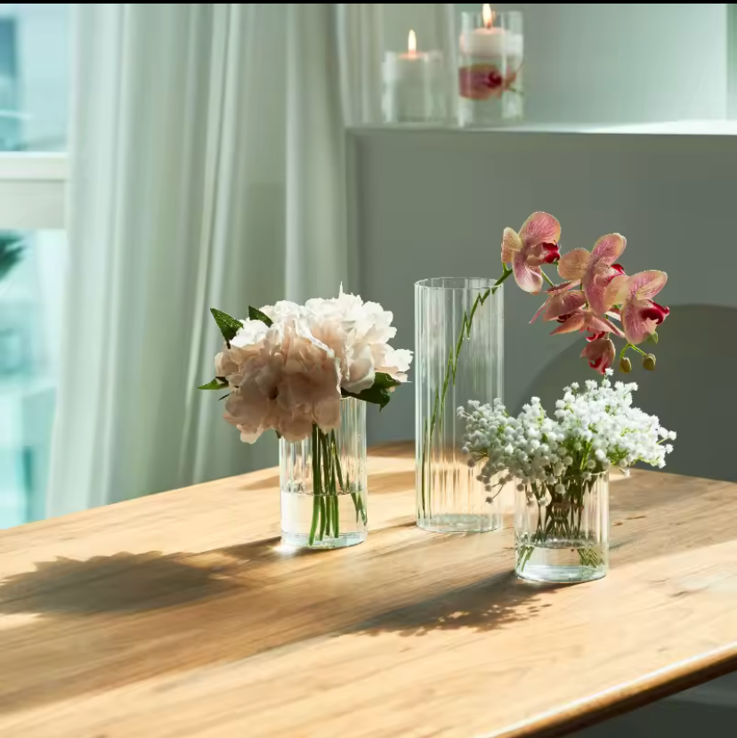 Three clear glass flower vases with pink, white, and green floral arrangements displayed on a wooden table in a bright room.