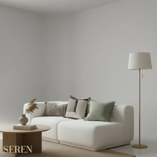Three neutral-toned decorative throw pillows arranged on a white sofa in a bright, minimalist room. One pillow in the foreground is a square taupe linen pillow with a solid border.